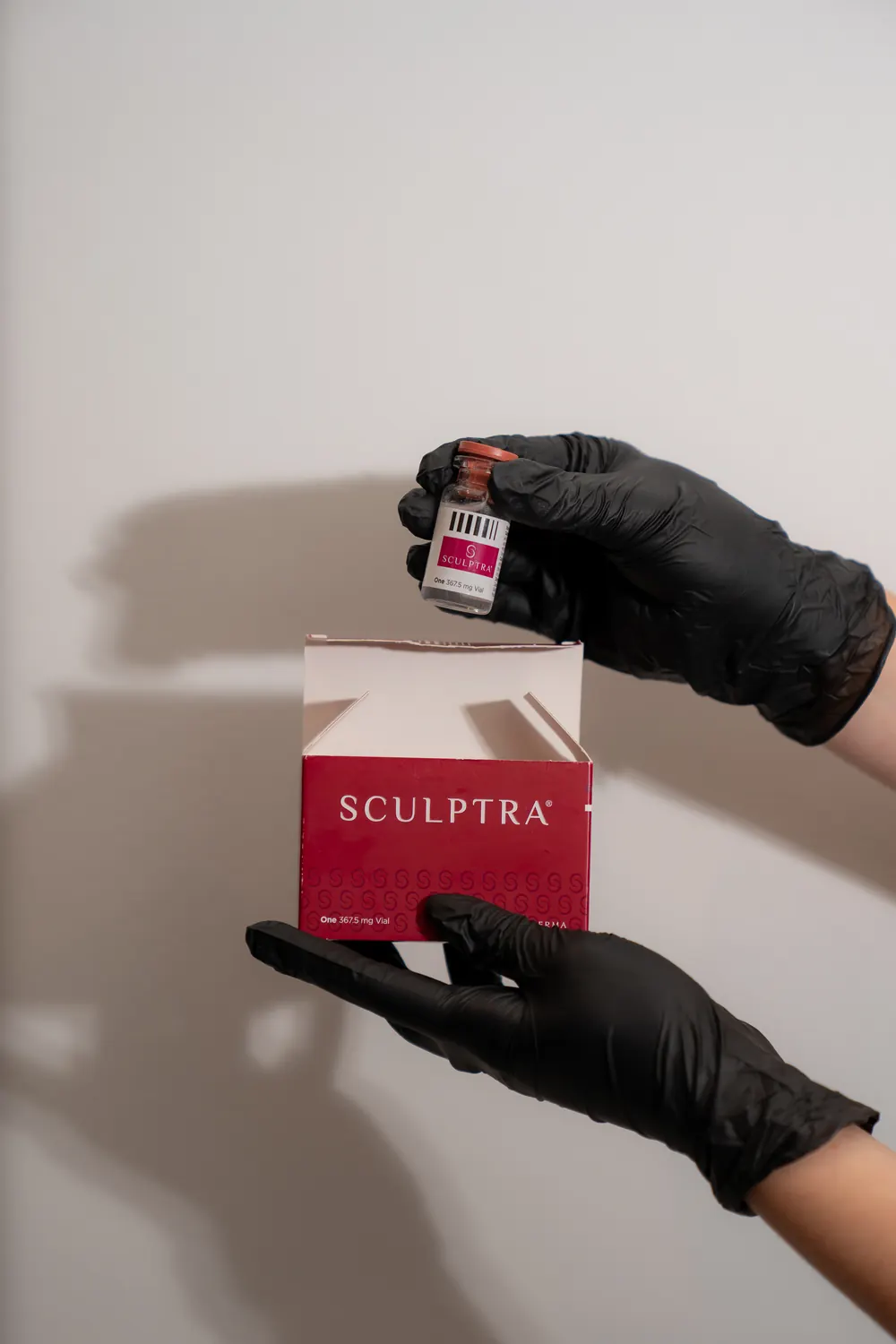 Gloved hands holding a vial and a box of Sculptra against a pale wall